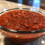 A close-up of thick, vibrant red homemade pizza sauce seasoned with herbs in a clear glass bowl.