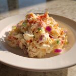 A scoop of creamy crab salad mixed with visible chunks of red onion and celery on a white plate.