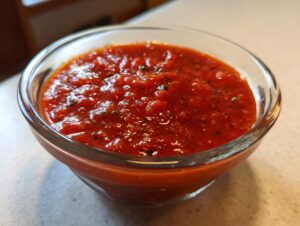 Close-up of rich, red, homemade pizza sauce seasoned with herbs in a clear glass bowl.