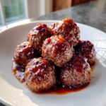 A pile of glossy, saucy cranberry meatballs glistening under warm light on a white plate.