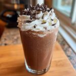 A tall glass of rich, blended frozen hot chocolate topped with whipped cream and chocolate shavings.
