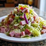A generous mound of chopped lettuce, cubed ham, red onion, and peppers forming a delicious grinder salad on a white plate.