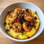 Close-up of creamy yellow shrimp and grits topped with seasoned shrimp, crumbled bacon, and fresh chives.