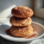 Three soft, thick Snickerdoodle cookies stacked vertically on a white plate, heavily coated in cinnamon sugar.