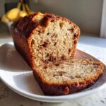 A close-up of a loaf of sugar free banana bread, sliced on a white plate, showing a moist interior.