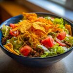 A close-up of a vibrant taco salad featuring ground beef, lettuce, tomatoes, shredded cheese, and tortilla chips in a dark blue bowl.