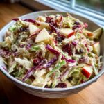 A close-up of a white bowl filled with creamy apple cranberry coleslaw, featuring shredded cabbage, apple chunks, and dried cranberries.