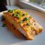 A close-up of a perfectly cooked fillet of bang bang salmon covered in creamy orange sauce and topped with fresh green onions.
