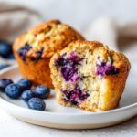 A blueberry protein muffins cut in half showing the moist crumb and burst blueberries inside, served on a white plate.