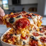 A hand lifts a cheesy, bubbling slice of pizza pasta bake topped with pepperoni and black olives.