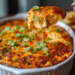 A spoonful of hot, cheesy, baked texas trash dip being lifted from a white casserole dish, showing cheese pull and green onion topping.