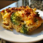 A close-up slice of creamy Chicken Divan casserole featuring pasta, chicken, broccoli florets, and a bubbly, browned cheddar cheese topping.