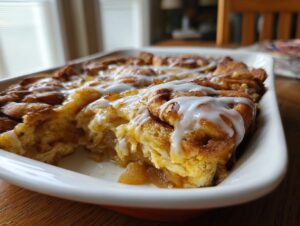 A baked cinnamon roll apple pie casserole topped with white icing, showing a serving removed.