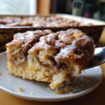 A fork lifts a gooey, glazed piece of cinnamon roll apple pie from a white plate.