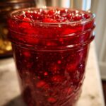 A close-up, vibrant shot of a glass jar filled with glistening, deep red christmas jam, showing whole and crushed fruit pieces.