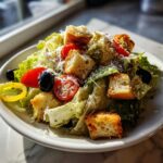 A close-up, sunlit photo of a bowl featuring a fresh copycat Olive Garden salad with romaine lettuce, cherry tomatoes, black olives, and large croutons.