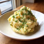 A generous serving of creamy garlic mashed potatoes topped with finely chopped green chives in a white bowl.
