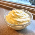 A close-up of thick, creamy, pale yellow homemade garlic sauce swirled high in a clear glass bowl.