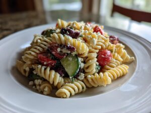 A serving of creamy mediterranean pasta salad featuring fusilli pasta, feta cheese, tomatoes, cucumbers, and olives on a white plate.