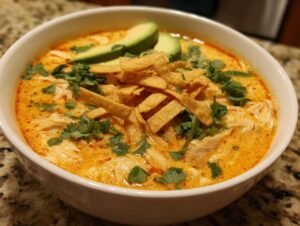 A close-up of a bowl of creamy, orange-hued tortilla soup recipe topped with shredded chicken, avocado, cilantro, and crispy tortilla strips.