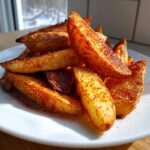 A pile of golden, crispy air fryer potato wedges heavily seasoned with red paprika on a white plate.