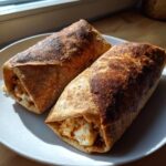 Two golden brown, crispy Baked Chicken Chimichangas resting on a white plate near a window.