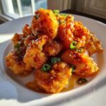 A close-up of crispy, saucy bang bang shrimp piled on a white plate and garnished with chopped green onions.