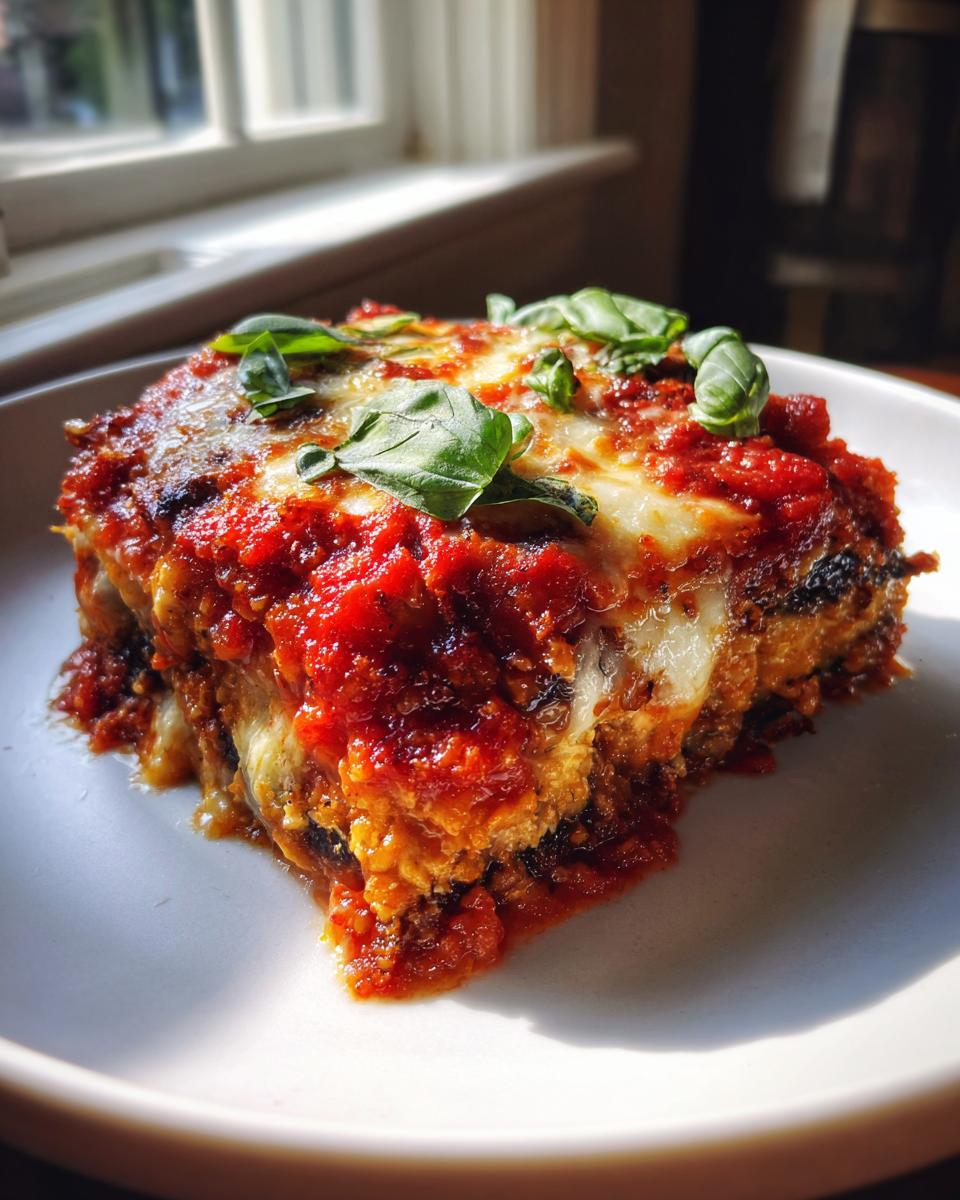 A close-up, sunlit serving of crispy eggplant parmesan topped with melted cheese, rich tomato sauce, and fresh basil leaves.