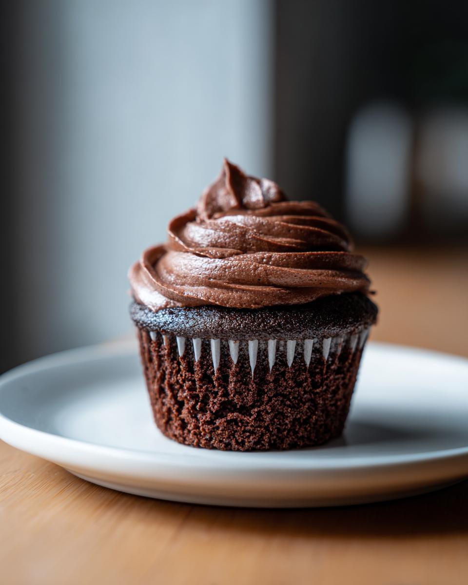 A single, rich, dark chocolate cupcake topped with swirled chocolate frosting, sitting on a white plate.