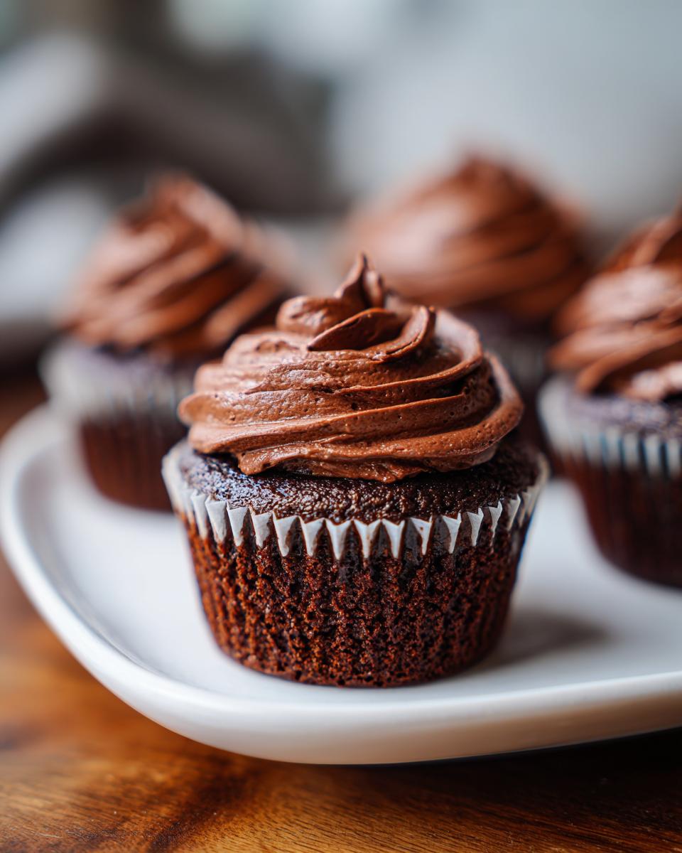 A close-up of one of the easy chocolate cupcakes topped with swirled, rich chocolate frosting, sitting on a white plate.