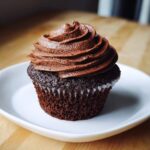 A single, rich, dark chocolate cupcake topped with a generous swirl of chocolate frosting, sitting on a white plate.