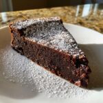 A rich, dark slice of flourless chocolate torte dusted generously with white powdered sugar on a white plate.