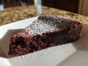 A decadent slice of flourless chocolate torte, dusted heavily with white powdered sugar, sitting on a white plate.