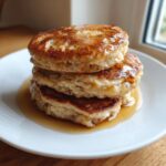 Close-up of a stack of three fluffy banana pancakes drizzled generously with maple syrup on a white plate.