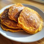 A stack of fluffy, golden-orange pumpkin pancakes dusted with powdered sugar on a white plate.