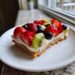 A close-up slice of fruit pizza sugar cookie topped with cream cheese frosting, strawberries, blueberries, and kiwi.