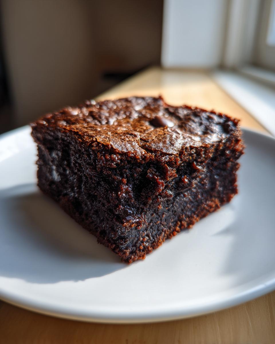 A rich, dark, fudgy square of sweet potato brownies sitting on a white plate in natural light.