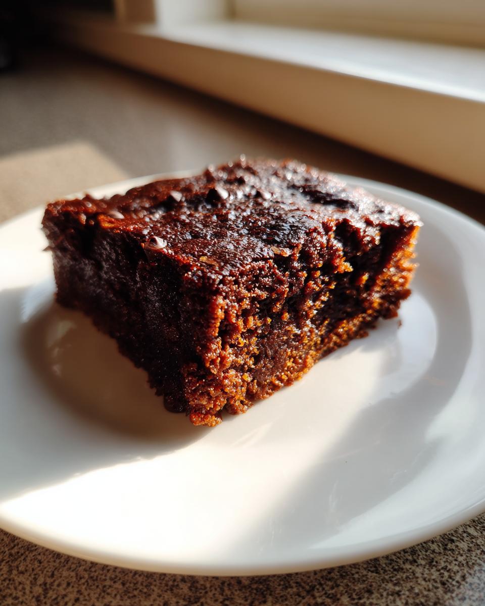 A close-up of a fudgy, dark brown sweet potato brownies square served on a white plate, highlighted by natural sunlight.