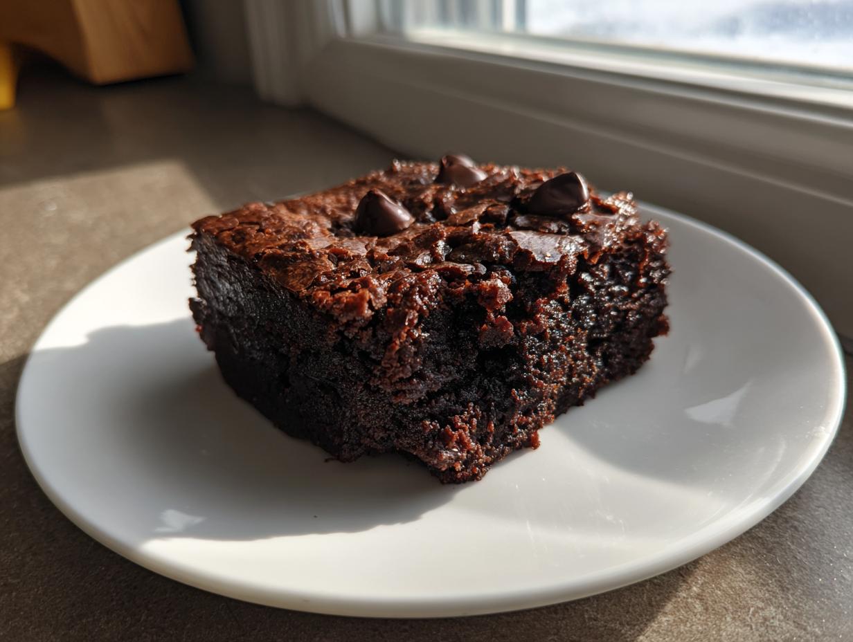 A single, fudgy square of dark chocolate sweet potato brownies topped with chocolate chips, sitting on a white plate.