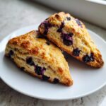 Two freshly baked, golden brown blueberry scones stacked slightly on a white plate.