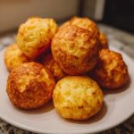 A close-up stack of freshly baked, golden-brown brazilian cheese bread bites on a white plate.