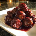 A close-up of a mound of savory Grape Jelly Meatballs coated in a thick, glossy, dark red sauce on a white plate.