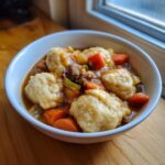 A close-up of a white bowl filled with rich ground beef and dumplings, featuring carrots and celery in a savory brown gravy.