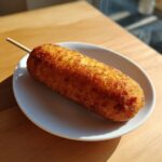Close-up of a perfectly fried, golden brown homemade corn dog on a stick, resting on a small white plate.