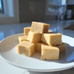 A stack of creamy, light brown peanut butter fudge recipe squares piled on a white plate in bright sunlight.