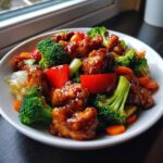 A close-up of glossy, saucy honey garlic chicken stir fry mixed with bright broccoli, carrots, and red bell peppers in a white bowl.
