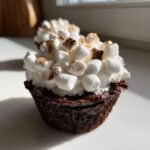 Close-up of a rich, dark hot chocolate cupcake topped with white frosting and toasted mini marshmallows.