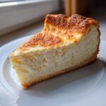 A close-up slice of rich, baked Keto cheesecake with a golden-brown top, resting on a white plate.