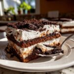 A decadent slice of layered Oreo dirt cake with cream and chocolate crumbs, being scooped by a silver spoon.