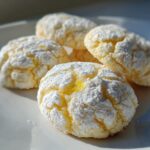 Close-up of several bright yellow lemon crinkle cookies heavily dusted with white powdered sugar on a white plate.